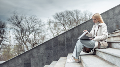 Young girl in a trench coat opens laptop while sitting on stairs