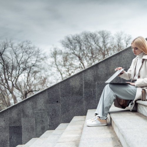 Young girl in a trench coat opens laptop while sitting on stairs